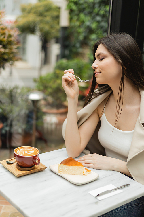 young woman with long hair holding fork near cheesecake next to cup of cappuccino on bistro table while sitting in leather jacket near window inside of modern cafe in Istanbul,stock image