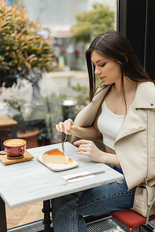 young  brunette woman holding fork near cheesecake next to cup of cappuccino with coffee art on bistro table while sitting in leather jacket near window inside of modern cafe in Istanbul,stock image