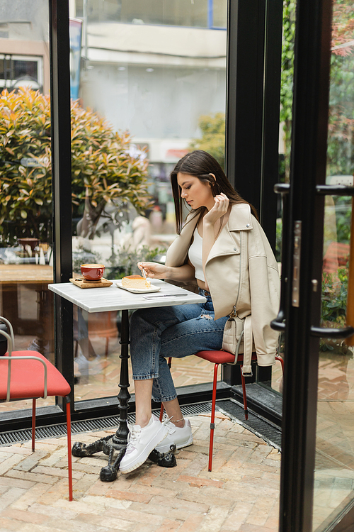 full length of young woman with long hair holding fork near cheesecake and cup of cappuccino on bistro table while sitting in leather jacket next window inside of modern cafe in Istanbul,stock image
