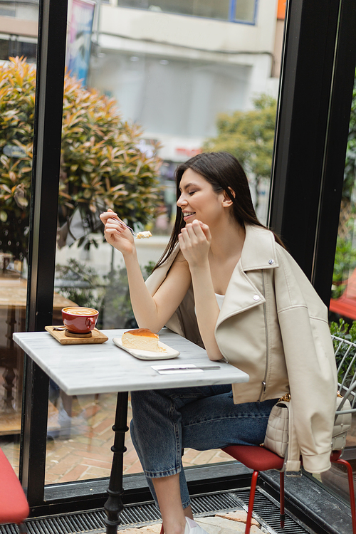 happy young woman with long hair eating cheesecake near cup of cappuccino with coffee art on bistro table while sitting in leather jacket next to window inside of modern cafe in Istanbul,stock image