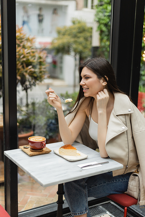 cheerful woman with long hair holding fork near cheesecake next to cup of cappuccino with coffee art on bistro table while sitting in leather jacket near window inside of modern cafe in Istanbul,stock image