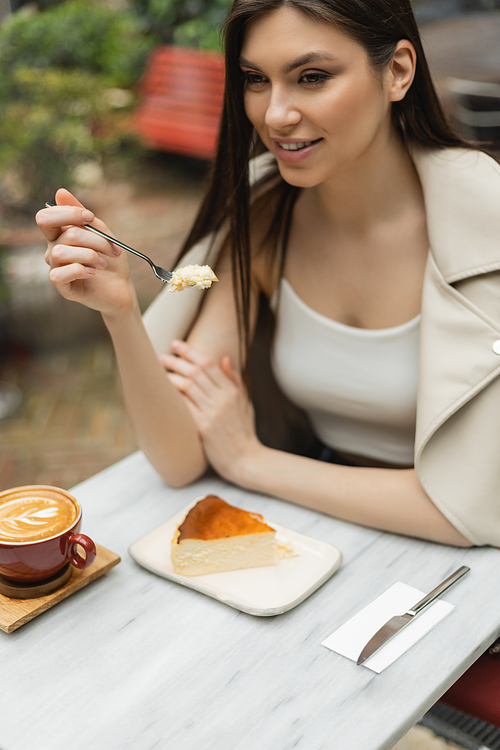 smiling woman with long hair holding fork with cheesecake next to cup of cappuccino with coffee art on bistro table while sitting inside of modern cafe in Istanbul,stock image