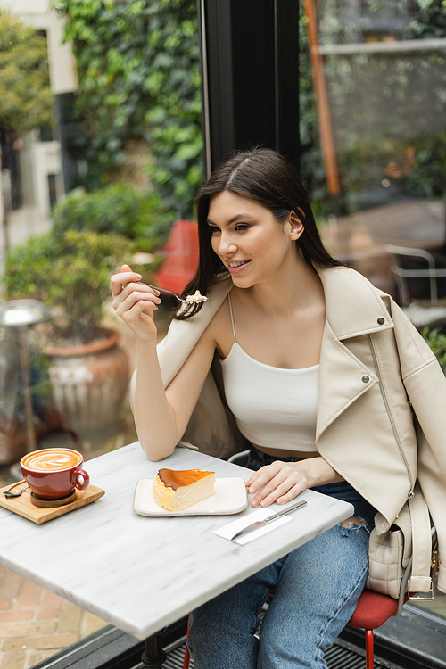 smiling woman with long hair holding fork near cheesecake next to cup of cappuccino with coffee art on bistro table while sitting next to window inside of modern cafe in Istanbul,stock image
