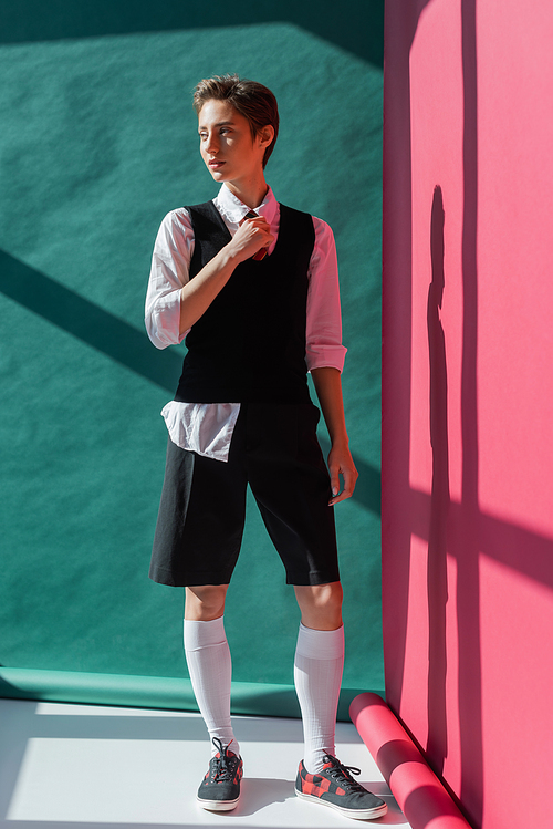 full length of stylish young woman with short hair posing in school uniform on pink and green