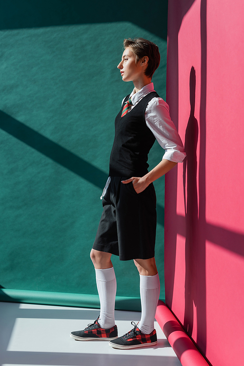 full length of stylish young student with short hair posing with hand in pocket of black shorts on pink and green
