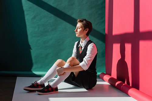 full length of stylish young woman with short hair sitting in school uniform on green and pink background
