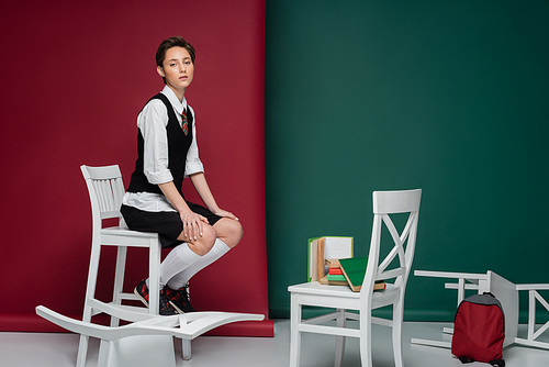 full length of stylish young woman with short hair sitting on chair near books on green and pink background
