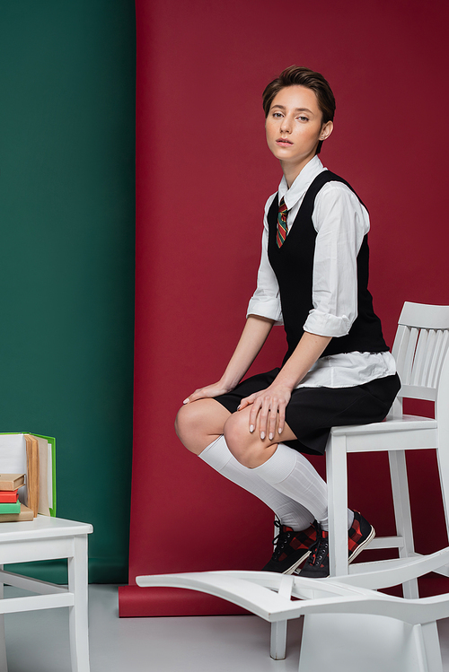 full length of stylish young student with short hair sitting on chair near books on green and red background