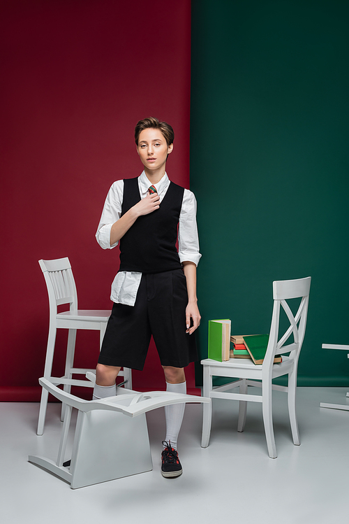 full length of stylish student with short hair holding book and standing near chairs on green and red background