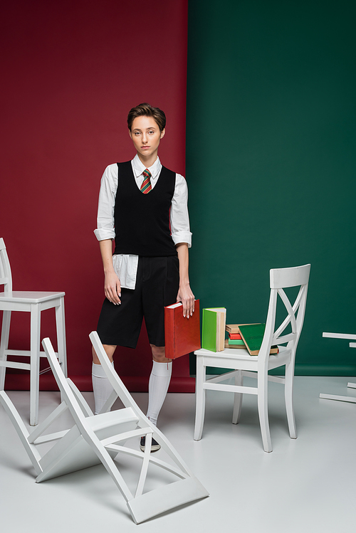 full length of stylish young woman with short hair holding book and standing near chairs on green and red background