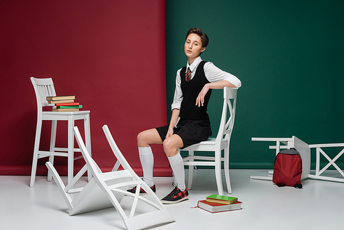full length of stylish young woman with short hair sitting on white chair among books on green and red background
