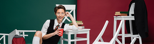 happy young student in eyeglasses holding drink in plastic cup near chairs and books on green and burgundy background, banner