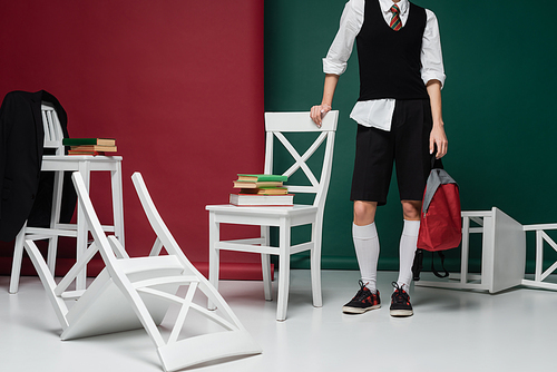 cropped view of stylish young student in school uniform standing around chairs with books on green and burgundy background