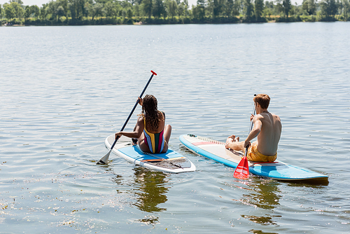 back view of brunette african american woman in striped swimsuit sailing on sup board near redhead and sportive man on picturesque lake during summer day