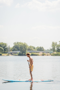 side view of young and redhead man in yellow swim shorts sailing on sup board with paddle on lake with green shore during water recreation on summer weekend