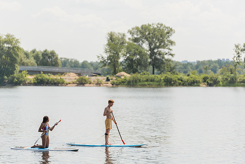 side view of young sportive man and african american woman standing on knees and sailing on sup boards with paddles on river with green bank in summer