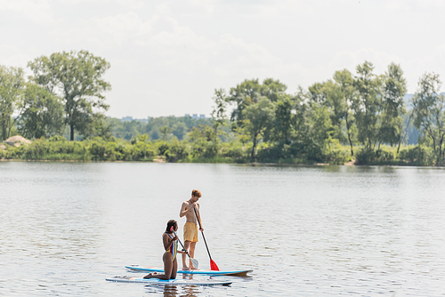 active multiethnic couple in colorful swimwear sailing on sup boards with paddles while spending summer vacation day on river with green trees on scenic bank