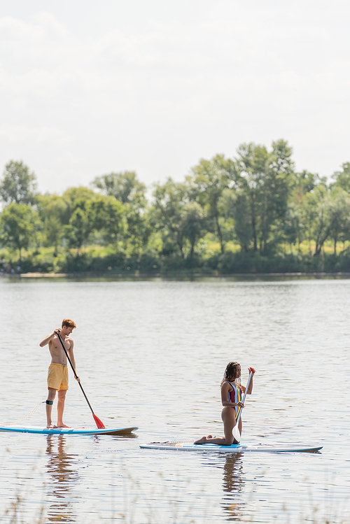 side view of young redhead man and african american woman in colorful swimsuit spending summer weekend while sailing on sup boards on picturesque river