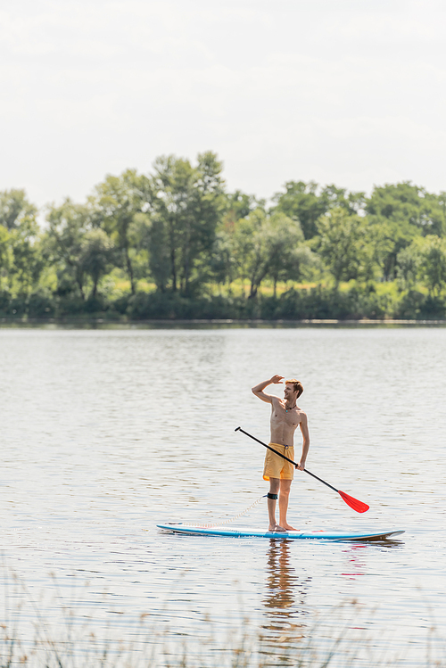 full length of sportive and redhead man in yellow swim shorts standing on sup board with paddle and looking away on lake with green trees on bank during summer weekend