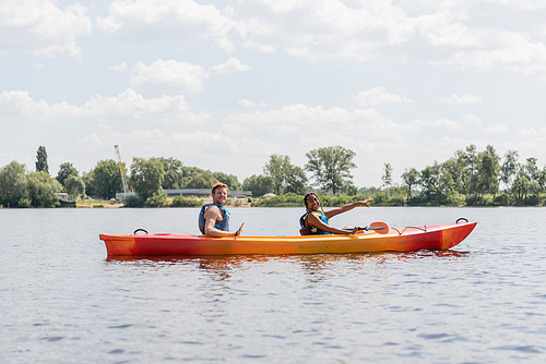 carefree african american woman in life vest pointing ahead and smiling at camera while sailing in sportive kayak with redhead friend during summer weekend on river