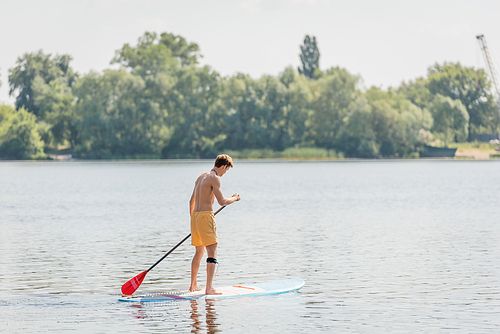 full length of redhead man in yellow swim shorts sailing on sup board with paddle while spending time on river with green picturesque bank on summer weekend