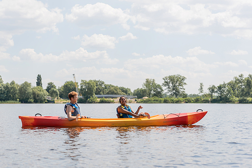 side view of joyful african american woman and young redhead man in life vests sailing in sportive kayak on river with green bank under blue and cloudy sky