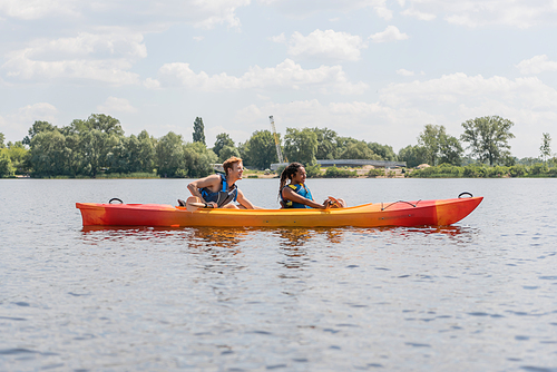 side view of brunette african american woman and young redhead man in life vests paddling in sportive kayak on lake with green shore under blue sky with clouds