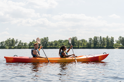 side view of smiling redhead man and brunette african american woman in life vests spending summer weekend by sailing in kayak on river under cloudy sky in summer