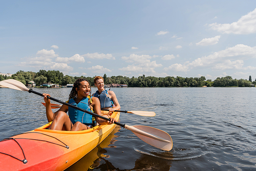 carefree and charming african american woman and young redhead man in life vests sailing in sportive kayak on calm water surface under blue sky with clouds on summer day