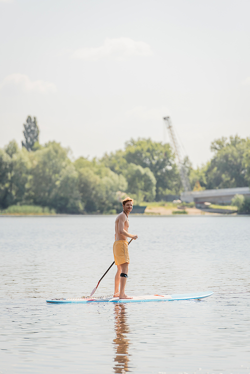 full length of sportive redhead man in yellow swim shorts holding paddle and looking at camera while sailing on picturesque lake on summer weekend day