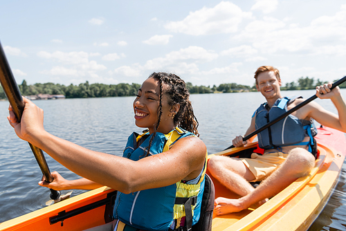 cheerful african american woman in life vest sailing in kayak while spending time on river with young and redhead man paddleboarding on blurred background