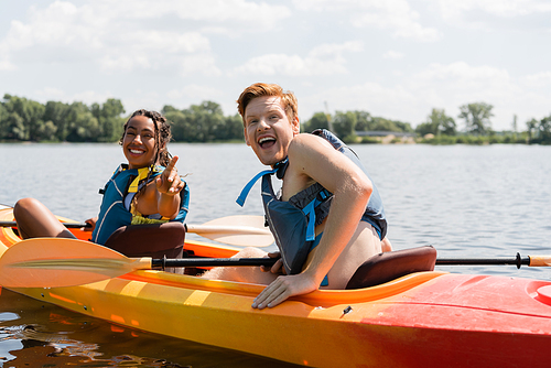 excited redhead man with open mouth looking at camera near cheerful african american woman in life vest pointing with finger while sailing in kayak on lake in summer