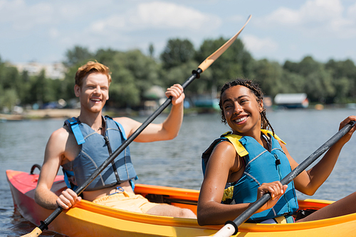 pleased african american woman and cheerful redhead man in life vests holding paddles while sailing in sportive kayak during summer weekend on lake on blurred background