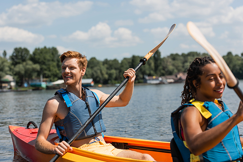 excited redhead man in life vest looking away while sailing in sportive kayak with pretty african american woman during recreation leisure on lake in summer