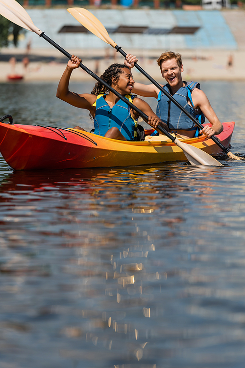 charming african american woman in life vest looking at excited redhead man while sailing in sportive kayak with paddles on summer day on blurred foreground