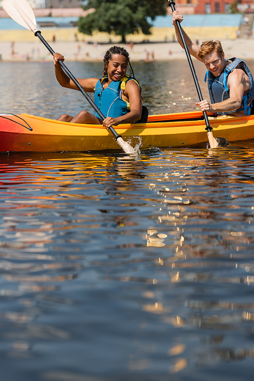 overjoyed african american woman and young redhead man in life vests looking at water splashes while paddling in sportive kayak during summer weekend