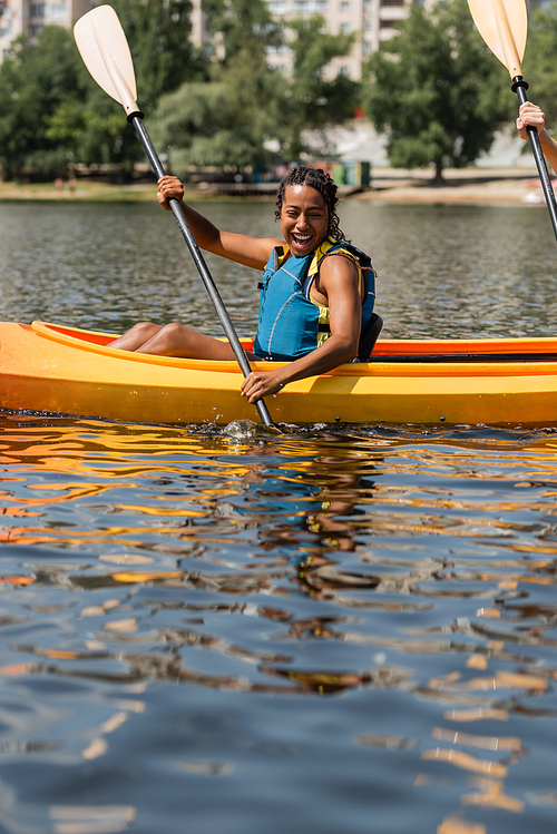 impressed and cheerful african american woman in life vest holding paddle while sailing in sportive kayak during recreation weekend on city lake on summer day