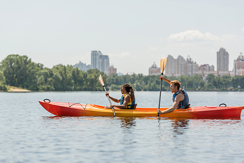 side view of active african american woman and excited redhead man in life vests paddling in sportive kayak along riverside with picturesque cityscape on blurred background