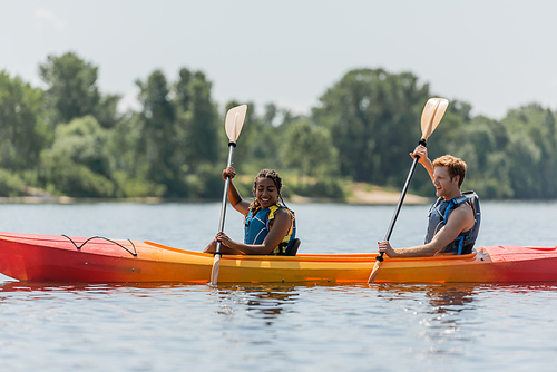 joyful and active interracial couple in life vests paddling in sportive kayak while spending time on lake with blurred green shore during summer vacation