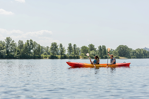 side view of african american woman with young man in life vests paddling in sportive kayak along green riverside during recreation activity in summer