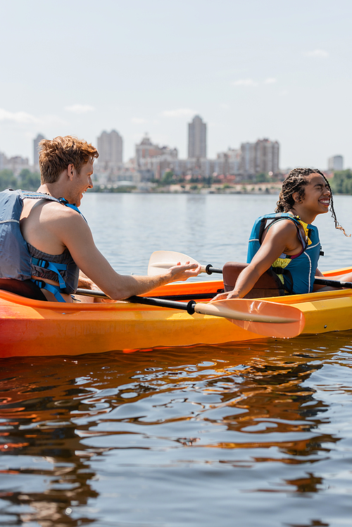 young redhead man in life vest talking to overjoyed african american woman laughing with closed eyes and sitting in sportive kayak on river with blurred cityscape on background