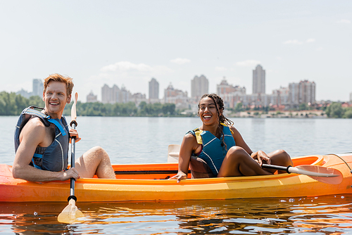 african american woman and redhead man in life vests sitting in sportive kayak with paddles and looking away on lake with picturesque cityscape on blurred background