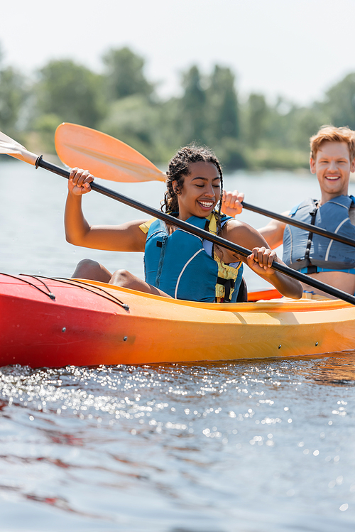 carefree and charming african american woman in life vest holding paddle and sailing in sportive kayak near young redhead man smiling on blurred background