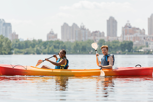 positive and sportive interracial couple in life vests sitting with paddles in kayak and smiling at camera on river with picturesque cityscape on blurred background