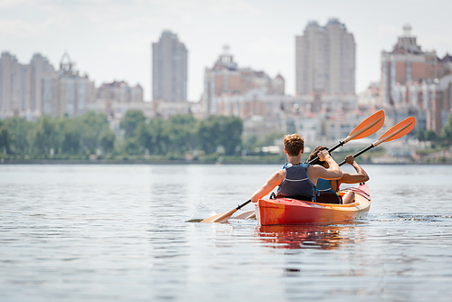 back view of active multiethnic couple in life vests paddling in sportive kayak on calm water surface on summer day with view of blurred urban buildings on riverside