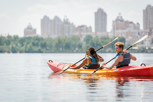 redhead man and african american woman in life vests spending time on river by sailing in sportive kayak and enjoying picturesque cityscape on blurred background