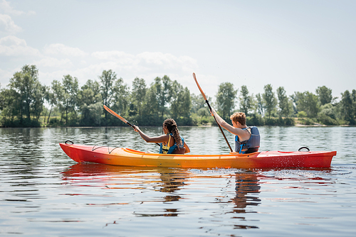 side view of active multiethnic couple in life vests holding paddles while sailing in sportive kayak on river with picturesque bank with green trees in summer