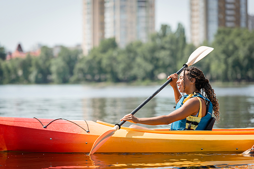 side view of active and charming african american woman in life vest sailing in sportive kayak with paddle on summer weekend day on blurred background