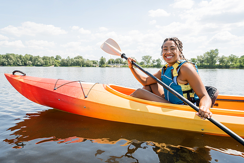 overjoyed african american woman in life vest paddling in sportive kayak and smiling at camera on lake with green picturesque shore on background in summer