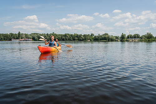 active multiethnic couple in safe vests sailing in sportive kayak on picturesque lake with green shore under blue and cloudy sky during water recreation on summer weekend